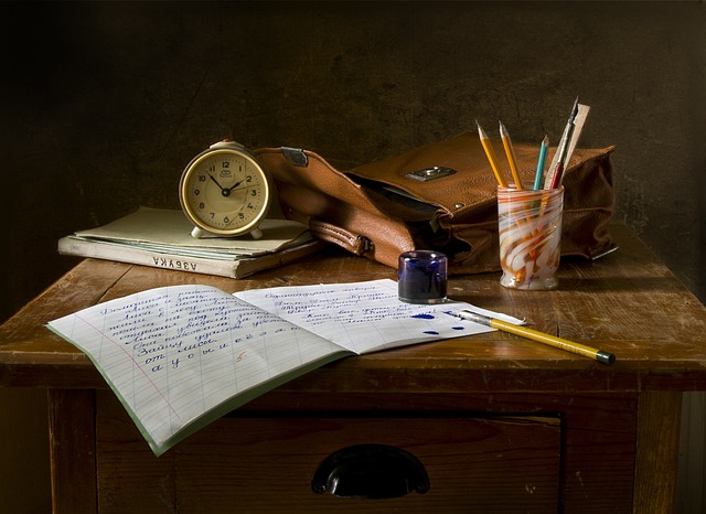 mesa com caderno aberto, bolsa, livro, despertador, pincel, tinta, lápis e régua dentro do copo