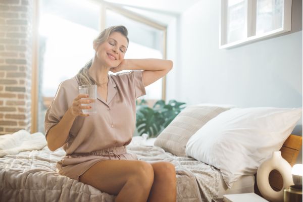 mulher sentada na cama com copo de agua, mão no pescoço e sorrindo
