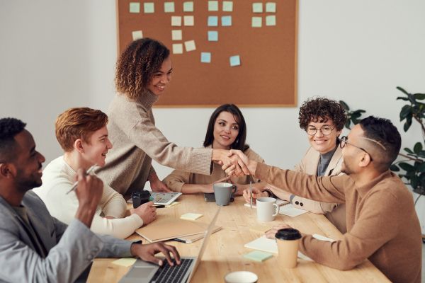 textos motivacionais trabalho em equipe pessoas sentada em volta de uma mesa com rapaz comprimentando uma moça em pé