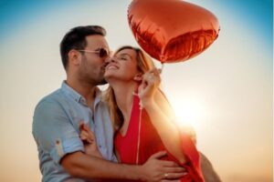 um homem beijando o rosto de uma mulher com balão de coração e raios de sol no fundo dos dois.