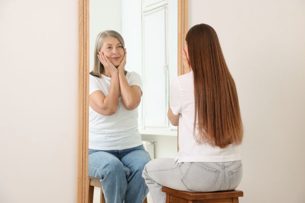 Mulher olhando no espelho e vendo uma versão mais jovem de si, simbolizando saudade de quem ela era.