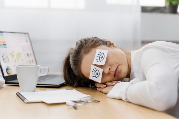 autosabotagem procrastinação no trabalho Pessoa dormindo na mesa de trabalho com papéis sobre os olhos, representando procrastinação e autossabotagem