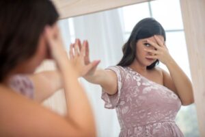 Mulher se olhando no espelho com expressão de insegurança e autocrítica, cobrindo parte do rosto